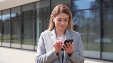 Modern cam binanın yanında dururken modern akıllı telefon kullanan resmi takım elbiseli genç bir iş kadını profesyonel bir bayan işveren dışarıda bir cep telefonuna internetten mesaj yazıyor..