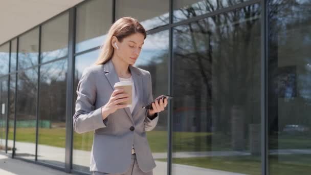 Cute attractive business woman using mobile phone app walking while walking holding coffee in urban city buildings look at smartphone screen feel happy smile at sunlight. Female stylish look. — Stock Video