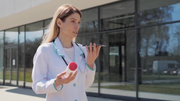 Kobieta lekarz trzymając butelkę pigułek i telefon komórkowy nagrywanie wiadomości głosowych online do pacjenta na zewnątrz podczas stania w pobliżu kliniki. Atrakcyjny lekarz trzymający butelkę leku w słoneczny dzień. — Wideo stockowe