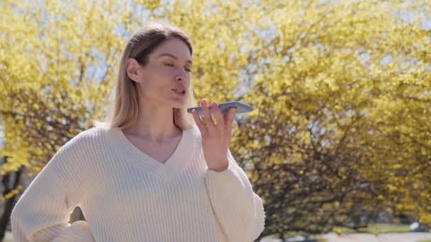 Красивая женщина в белом свитере с помощью записи мобильного телефона отправляет голосовое аудио сообщение на громкоговорителе в парке рядом с деревом с желтыми листьями в теплый солнечный день цифровой мобильной помощи. — стоковое видео