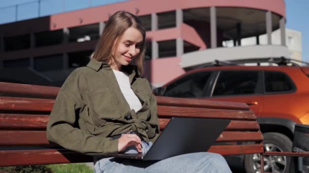 Young beautiful girl freelancer works at a laptop sitting on a wooden bench smiling having good mood in the rays of the sunset. Female person working outside the office in the open air near parking. — ストック動画