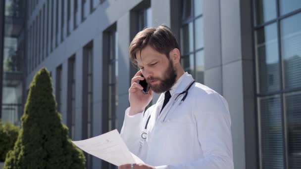 Confident therapist giving consultation by phone remotely, standing outdoors. Professional male doctor holds a report on patients illness gives advice patient on the background of modern clinic. — Stock Video