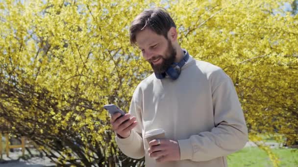 Przystojniak korzystający ze smartfona na ulicy. Przystojny Mężczyzna noszący luźne ubrania, słuchawki trzymające kawę podczas pisania w telefonie komórkowym przewijania mediów społecznościowych. Internetowa komunikacja internetowa. — Wideo stockowe