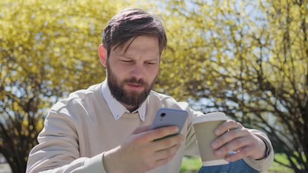Handsome happy bearded man sitting on the curb with cup of coffee using mobile phone browsing internet texting message on social media dating app having free time some rest for communication with girl — Stock video