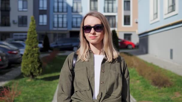 Girl wide smiling with healthy white teeth. Beautiful blonde girl adjusts sunglasses with her hand and smiles at the camera on the background of city buildings. Portrait of pretty female face model. — Vídeo de stock