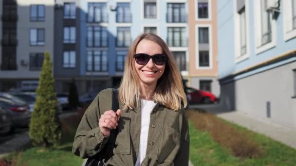 Portrait of young attractive smiling woman in sunglasses looking at camera on a background of colored city buildings. Portrait happy female tourist having good time wandering around town in summer. — 비디오