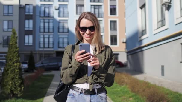 Portrait of smiling young woman wearing sunglasses using mobile phone typing message browsing internet social media outside in the city on sunny day. Satisfied female Internet user enjoy conversation. — Vídeos de Stock