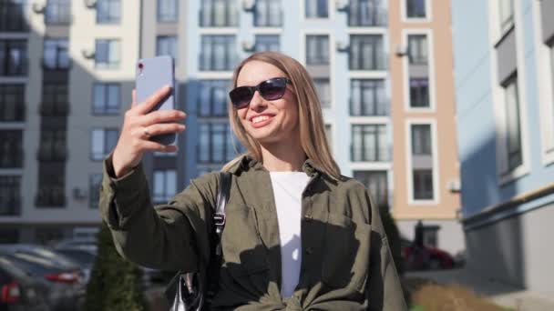 Beautiful Happy Blonde Girl Wearing Sunglasses Making Online Video call Smiling Talking Waving her Hand to Friend Enjoys the Conversation Walking Relaxed on the Street City Background Outdoor. — Stockvideo