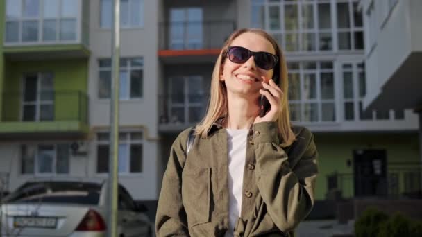 Boldog fehér nő hív telefonon naplementekor. Portré vonzó vidám lány beszél okostelefonon közelében zöld városi épületek. Aranyos hölgy használja a modern modult a kommunikációhoz. Nyári idő. — Stock videók