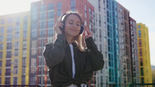 Szczęśliwa wesoła dziewczyna słuchająca muzyki pokazującej kciuk w tle miejskiego miasta. Młoda kobieta tańcząca w centrum emocjonalna blondynka zdejmuje słuchawki i uśmiecha się szczerze na zewnątrz. — Wideo stockowe
