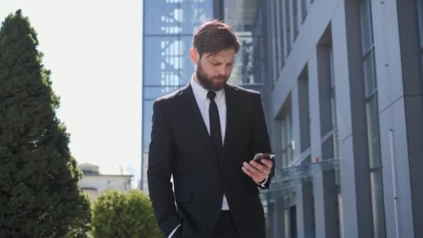 Молодой уверенный в себе бизнесмен в современном костюме идет к себе в офис и использует мобильный телефон в чате с коллегами на фоне бизнес-здания. СМС для мужчин на открытом воздухе. — стоковое видео