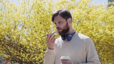 Erkek sakallı hippi elinde akıllı telefonuyla mikrofonla konuşuyor. Elinde kahve olan sesli mesaj gönderiyor. Parktaki sarı ağaçların arka planında cep telefonundan sesli mesajlar kaydeden yakışıklı bir adam..