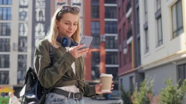 Cheerful young woman in sunglasses wireless headphones and coffee standing in the city while using mobile phone. Messaging browsing internet, social media. Girl having happy smile, stylish clothes.