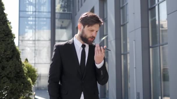Stylish bearded man making voice message or using virtual assistant app on mobile phone. Confident businessman recording voice message on speakerphone while standing near modern office building. — Stockvideo