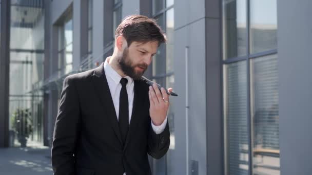 Bearded business man making voice message using online virtual assistant app looking confident. Handsome male banker talking and using smartphone while standing at street in office district outside. — Stockvideo