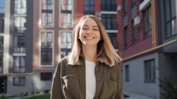 Cute blonde woman walking on the urban street looking at camera. Portrait of beautiful happy girl with healthy white teeth standing in city near beautiful colored buildings. Positive emotion. Freedom. — Stockvideo