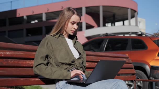 Concentrated young beautiful girl freelancer works at a laptop sitting on a wooden bench in the rays of the sunset. Female person working outside the office in the open air near parking. Messaging. — Video Stock