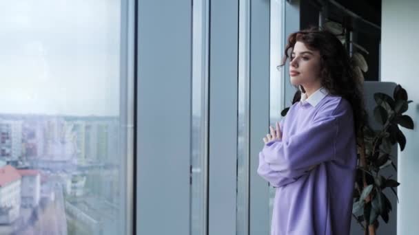 Beautiful thoughtful woman freelancer coming to the window. Pretty girl with deep brown eyes standing in office near panoramic window with a city landscape. The talented designer is inspired. — Vídeo de Stock