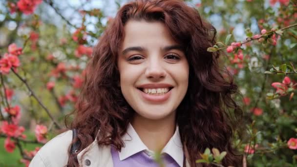 Portrait of a beautiful girl in a purple sweater with curly hair standing near a tree with spring flowers. Woman smiling and enjoying the fresh air, having good mood and looking into the camera. — Vídeo de stock