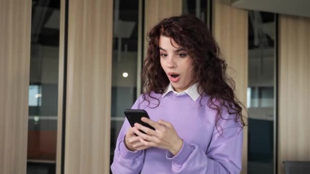 Overjoyed attractive woman freelancer celebrate online win, success hearing great news, feel amazed. Beautiful girl with curly hair deep brown eyes reading good news on her phone in the office. — Vídeos de Stock