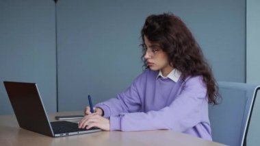 Young business woman freelancer typing on laptop making notes in a notebook working in internet, beautiful female professional designer in glasses using pc technology doing online job in office.