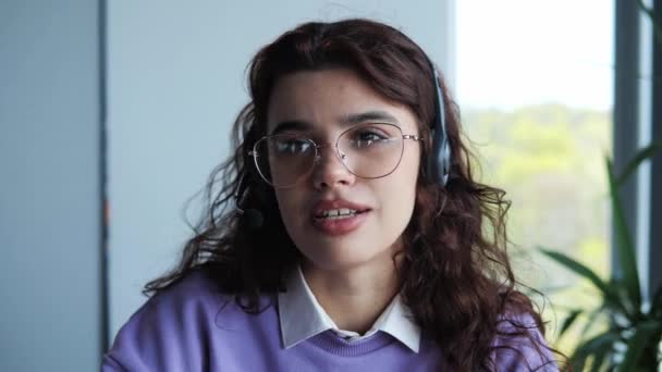 Cheerful student girl wears headphones and glasses, studies online with an Internet teacher, learns a language, talks, looks at a laptop, focuses on video calls, distance job, learning concept. — Vídeo de Stock