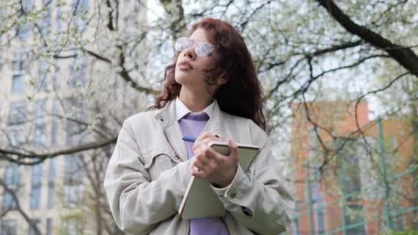 Cheerful thoughtful girl with curly hair making notes in notebook. Smiling female designer enjoying nature standing near beautiful tree. Remote work and freelancer Lifestyle Concept. — Vídeos de Stock