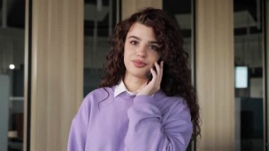 Kreatif ofiste cep telefonuyla konuşan kıvırcık saçlı ve mor kazaklı mutlu bir kadın. Portre gülümseyen kadın mimar ev telefonuyla arıyor. İş adamları fikirler tasarlar.