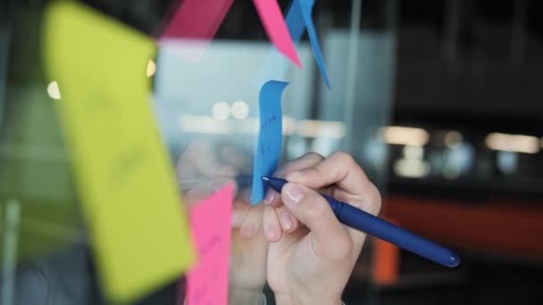 Close up view of female hands manager writing notes on colorful paper stickers, glass board in office. Girl working in modern office, making planning. Woman hands writing reminder on blue sticker. — Vídeo de Stock