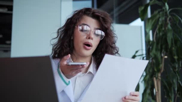 Young beautiful curly hair female doctor in lab coat and glasses recording audio message, reading conclusion of the disease talking on mobile phone while working at desk in medical office. — ストック動画