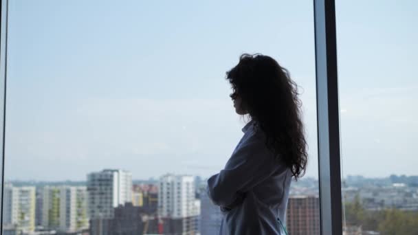Portrait of attractive sad woman doctor with curly hair looking through the window city background and feeling tired. Thoughtful silhouette of an intern female doctor, thinking face by the window. — Stock Video