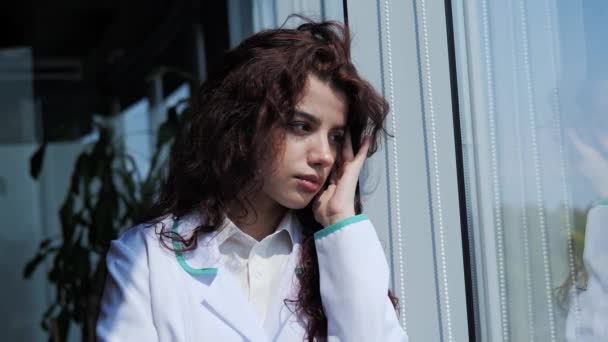 Close up view of sad tired woman doctor with curly hair looking through the window city background and feeling tired. Thoughtful silhouette of an intern female doctor, thinking face Headache. — Stock video