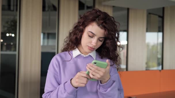 Beautiful female freelancer with curly hair typing message on mobile phone in office. Young girl designer in a purple sweater chatting on phone. Close up young woman hands using smartphone indoor. — Vídeos de Stock