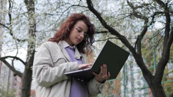 Young redhead girl with curly hair using laptop new technology internet connection standing by a tree in spring day. Distance learning, online education, work outside. Student communicate remotely. — Stockvideo