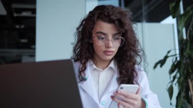 Laboratuvar önlüğü ve gözlüklü, cep telefonu ve dizüstü bilgisayarla çalışan odaklanmış kıvırcık saçlı kadın doktor, hastalığın sonucunu okuyarak tıp bürosundaki masada oturan bilgisayara giriyor..