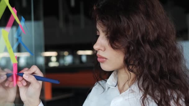 Focused smart office worker girl writing on stickers on glass board in modern workplace. Closeup concentrated business woman searching for proper solution indoors. Pretty girl working with interest. — Stockvideo