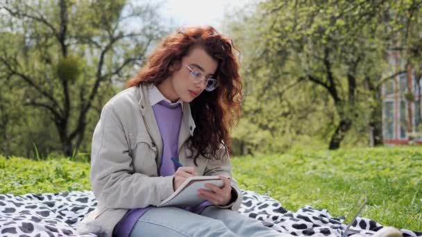 Charming girl with curly hair in a purple sweater sits on the grass and makes notes in a notebook. Girl freelancer doing remotely work enjoying fresh spring air in the garden. — Vídeos de Stock