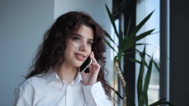 Pretty young girl standing near the window in the office while talking on the mobile phone. Female entrepreneur with curly hair deep brown eyes dressed in white shirt having call conversation indoor. — ストック動画