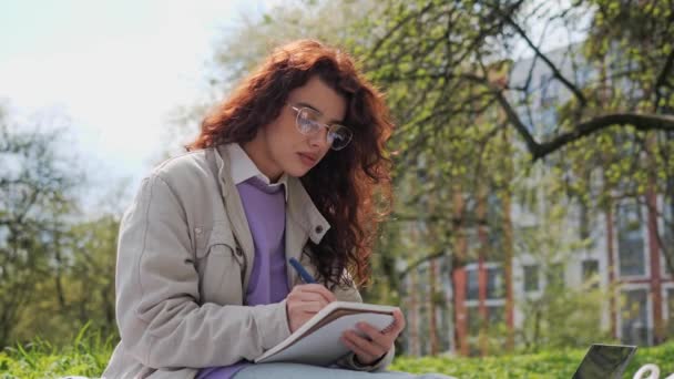 Pretty student with curly hair in a purple sweater sits on the grass and makes notes in a notebook. Girl freelancer doing remotely work enjoying fresh spring air in the garden. Distance learning. — Video Stock