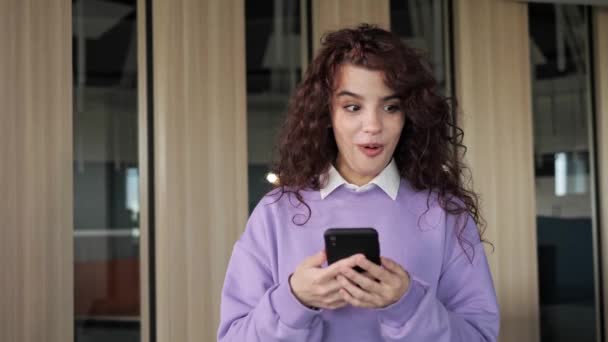 Excited attractive woman freelancer celebrate online win, success hearing great news, feel amazed. Beautiful girl with curly hair deep brown eyes reading good news on her phone in the office. — Vídeos de Stock