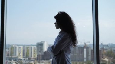 Dalgın, kıvırcık saçlı kadın doktor pencere arkasını inceliyor ve düşünüyor. Stajyer bir kadın doktorun düşünceli silueti. Klinikte dikilirken pencereden dışarı bakan kız..