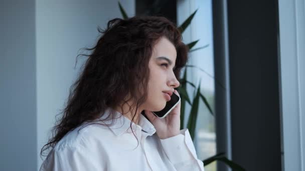 Όμορφο κορίτσι στέκεται κοντά στο παράθυρο του γραφείου, ενώ μιλάει στο κινητό τηλέφωνο. Γυναίκα επιχειρηματίας με σγουρά μαλλιά βαθιά καστανά μάτια ντυμένος με λευκό πουκάμισο έχοντας τηλεφωνική συνομιλία εσωτερική. — Αρχείο Βίντεο