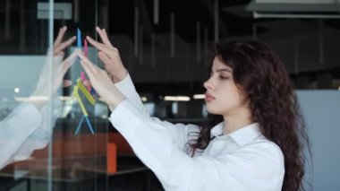 Güzel bir ofis çalışanı, farklı görevler için yapıştırma talimatları yapıştırır, şeffaf duvara yapıştırılmış yapışkan notlar kullanarak yeni projeler oluşturur. Hedefleri böl, önemli hatırlatıcılar kavramı oluştur.