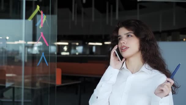 Female office worker talking on mobile phone standing near glass wall in office, strategizing new projects using sticky notes placed on transparent wall. Subdivide goals, make important reminders. — Video Stock