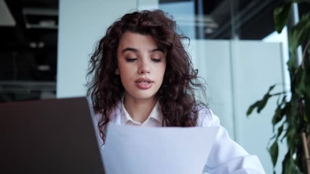 Young beautiful curly hair female doctor woman surgeon practitioner wears medical coat, reading conclusion of the disease consulting patient remote using laptop online video conferencing in clinic. — Video