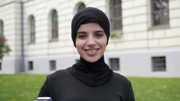 Портрет красивої молодої студентки мусульманської дівчини в чорному традиційному хіджабі, дивлячись прямо на камеру і посміхаючись. Крупним планом жіноче красиве обличчя з чарівною посмішкою, що стоїть за межами університету . — стокове відео