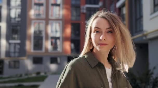 Beautiful blonde girl with blue eyes walks down the street turns to the camera and smiles. Female woman with natural beauty turning to camera having fun looking great straightens hair outdoors. — Stockvideo