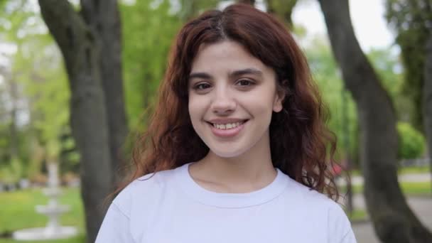 Portrait of curly hair happy girl outdoor. Girl face in park close-up. Dream girl. Happy face with charming smile standing outside. Female model wearing white t-shirt has a beautiful smile. — Stockvideo