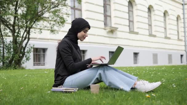 Elegant muslim arab islamic student girl in hijab. Woman freelancer user uses laptop sitting on green grass in park near university working online studying remotely chatting browsing shopping outdoor. — Wideo stockowe