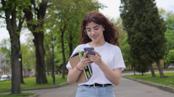 Cheerful curly hair student in white t-shirt with books walking near university using mobile phone in the park outdoor. Beautiful girl surfing social media, internet. Lifestyle and education concept. — Vídeos de Stock